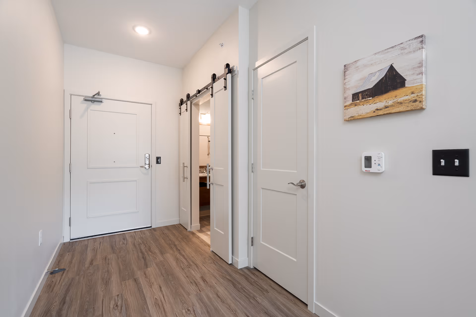 Interior view of a hallway in a senior living facility with a white entrance door, a sliding barn door leading to a bathroom, a closed white door, wood flooring, a thermostat on the wall, and a framed picture of a barn hanging above the thermostat.