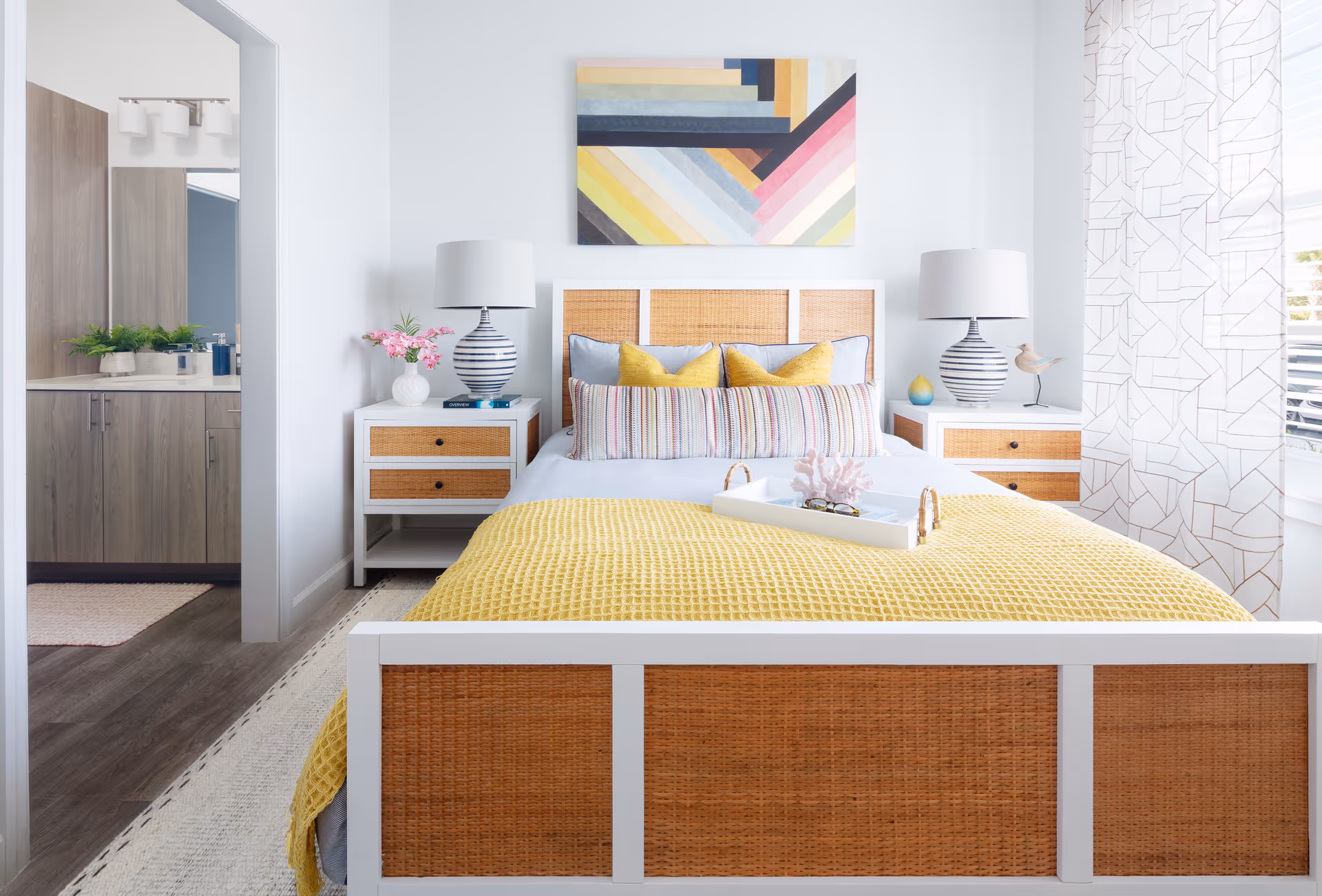 A bright and airy bedroom with a bed featuring a white frame and woven rattan panels. The bed is made with light blue sheets, a yellow textured blanket, and multiple pillows in yellow and striped patterns. On either side of the bed are white nightstands with matching woven rattan drawers, each topped with a striped lamp and decorative items. A colorful geometric painting hangs above the bed. To the left, an open doorway reveals a bathroom with wooden cabinetry and a large mirror. Light filters in through a window with sheer patterned curtains on the right side of the room.