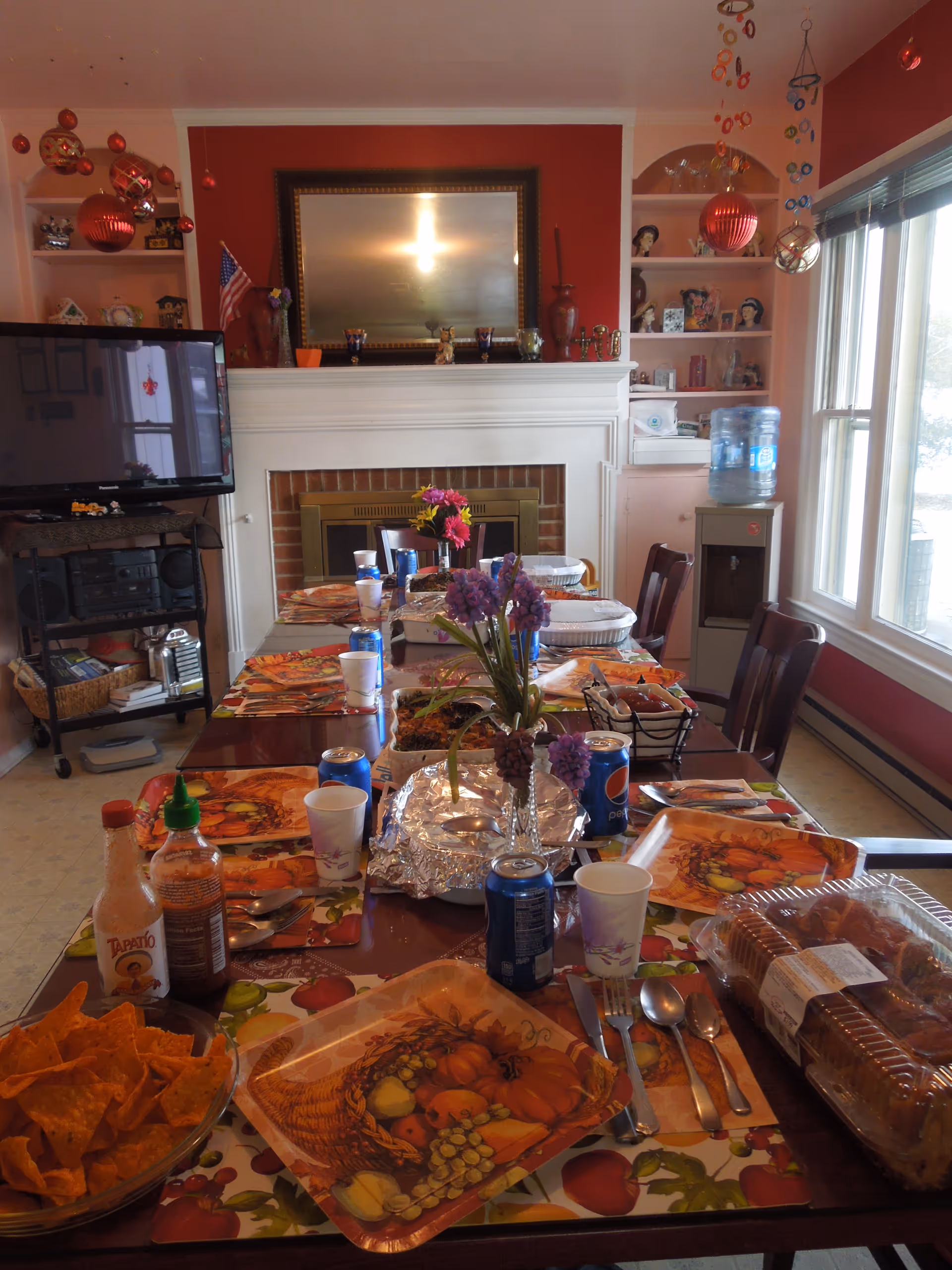 A dining room set for a meal with a long table covered with autumn-themed placemats and plates. The table has various food items, drinks including cans of Pepsi, and condiments like Tapatio hot sauce. There are flowers in vases as centerpieces. In the background, there is a fireplace with a large mirror above it, shelves with decorations, a television on a stand, and a water dispenser near a window letting in natural light.