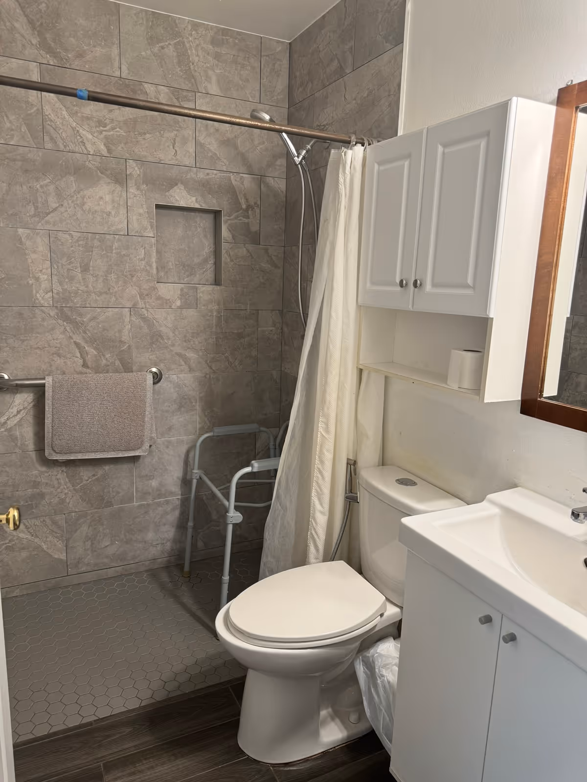 Small accessible bathroom with a tiled walk-in shower, shower chair and grab bar, toilet, sink vanity, and wall cabinet.
