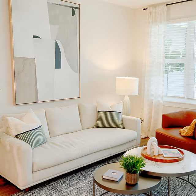 A bright living room with a white sofa adorned with white and green striped pillows, a modern abstract painting on the wall above the sofa, a round coffee table with a small green plant and a book, a white lamp on a side table, sheer curtains on the window, and a brown armchair with a yellow pillow.