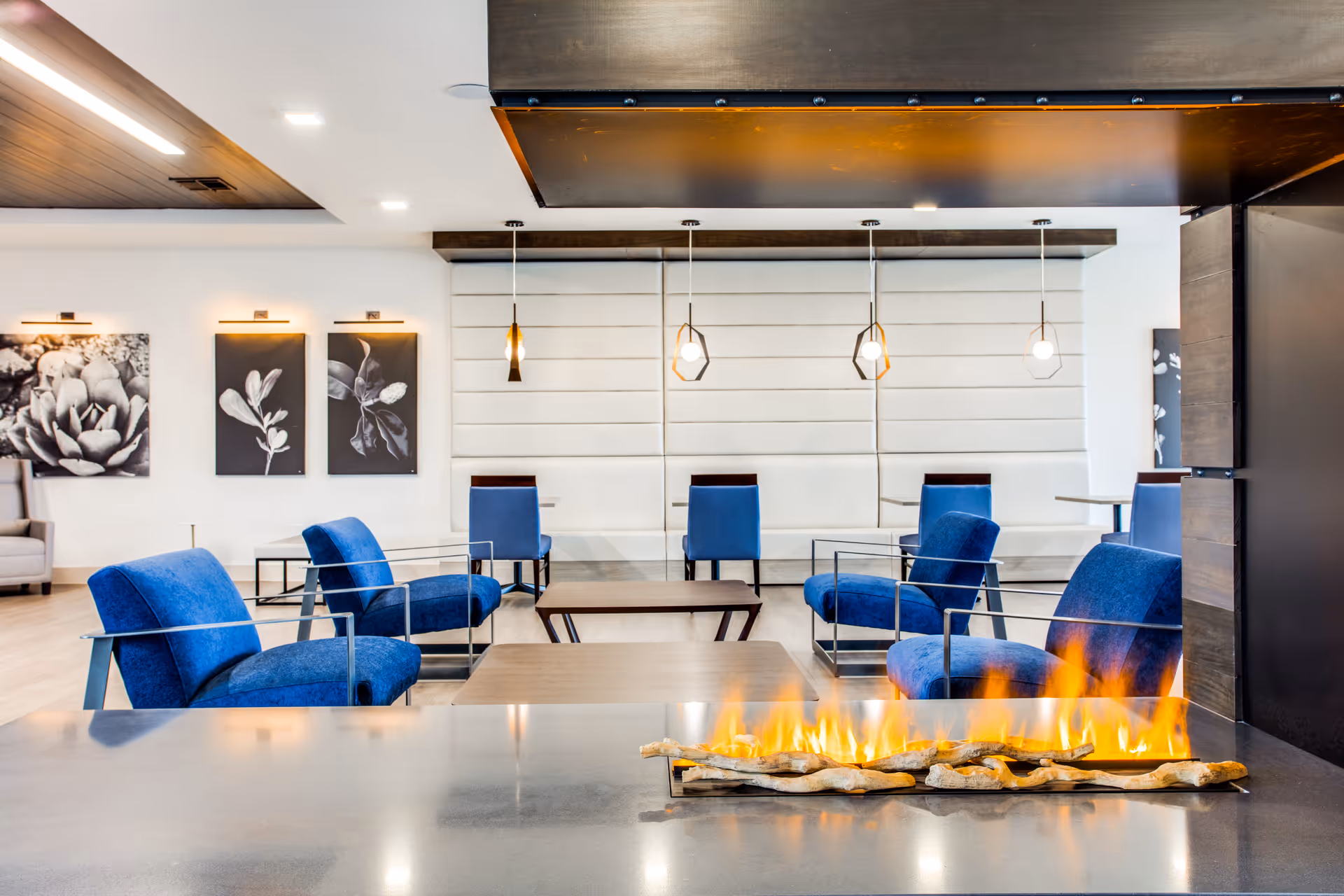 Modern lounge area with a row of blue armchairs and a central coffee table, featuring a contemporary linear gas fireplace in the foreground. The room has white paneled walls, hanging pendant lights, and black and white botanical artwork on the walls.