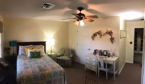 A cozy bedroom with a bed covered in a colorful patterned quilt, a bedside table with a lamp, a patterned armchair, a white desk with a chair, books, and a coffee maker. The room has beige walls, a ceiling fan with lights, and decorative floral wall hangings above the desk.