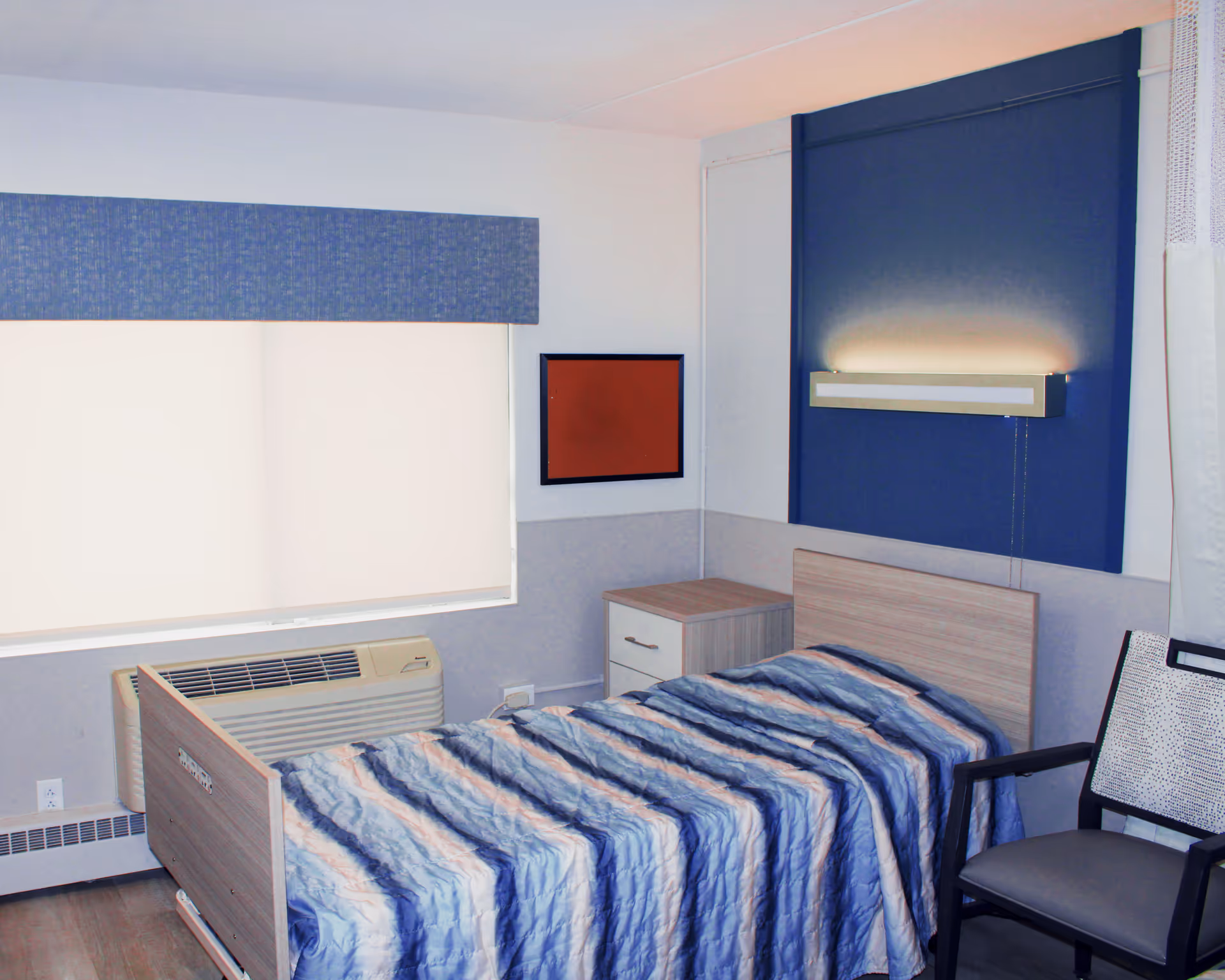 A single bed with a blue and white striped blanket in a small room. There is a wooden nightstand next to the bed, a chair with a patterned backrest, a window with a blue valance and closed blinds, and a wall-mounted light fixture above the bed on a blue accent panel.