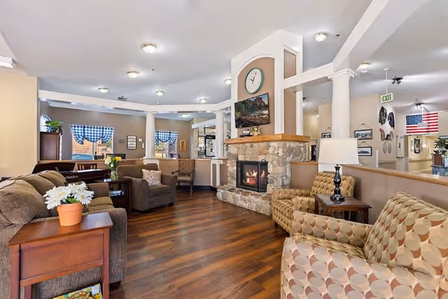 A cozy living room area in a senior living facility with wooden flooring, a stone fireplace with a clock and TV above it, several upholstered chairs and sofas, a side table with a lamp, and windows with blue checkered curtains. The room is well-lit with ceiling lights and has decorative columns and artwork on the walls.