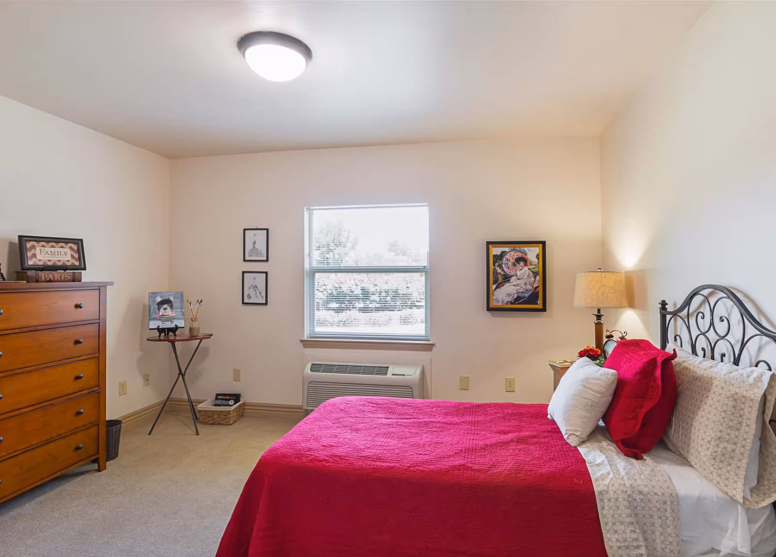 A bedroom in Bethesda Gardens Memory Care Community featuring a bed with a red quilt and multiple pillows, a wooden dresser with decorative items on top, a small table with art supplies and a painting of a dog, framed artwork on the walls, a window with blinds, and a lamp on a bedside table.