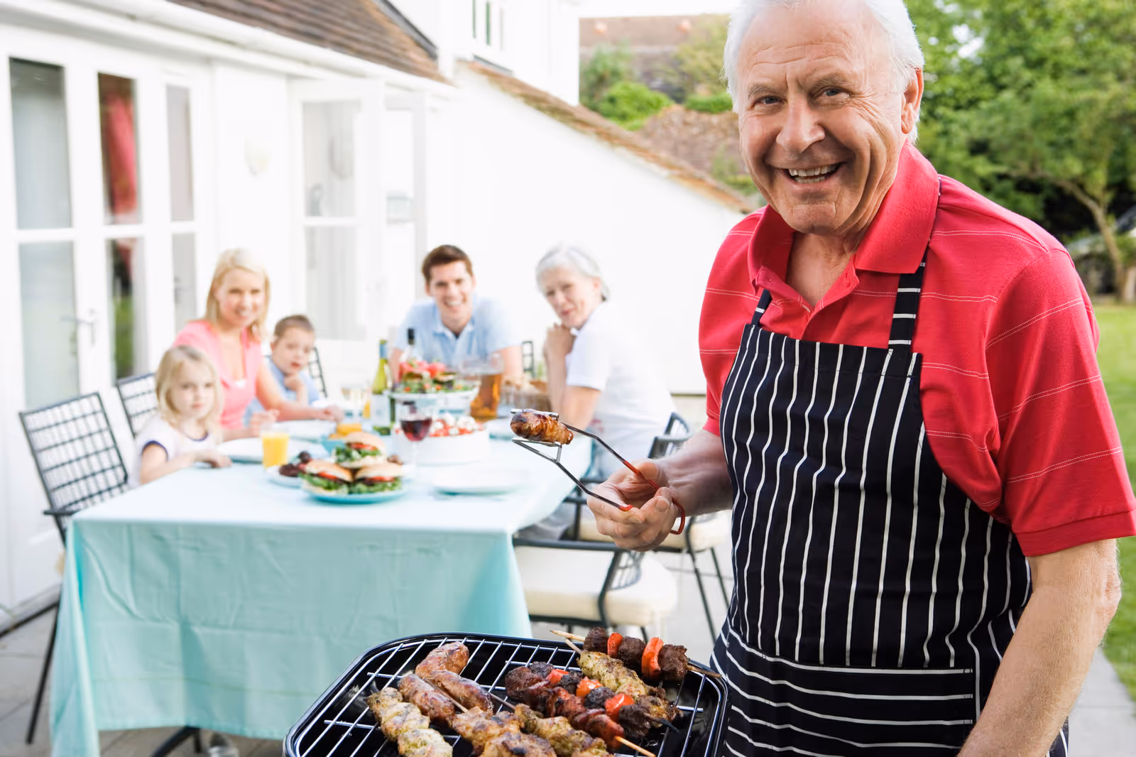 An elderly man wearing a red polo shirt and a black and white striped apron is smiling while grilling skewers of meat and vegetables outdoors. In the background, a family of five, including two children and three adults, are seated at a table covered with a light blue tablecloth, enjoying a meal together outside a white house.