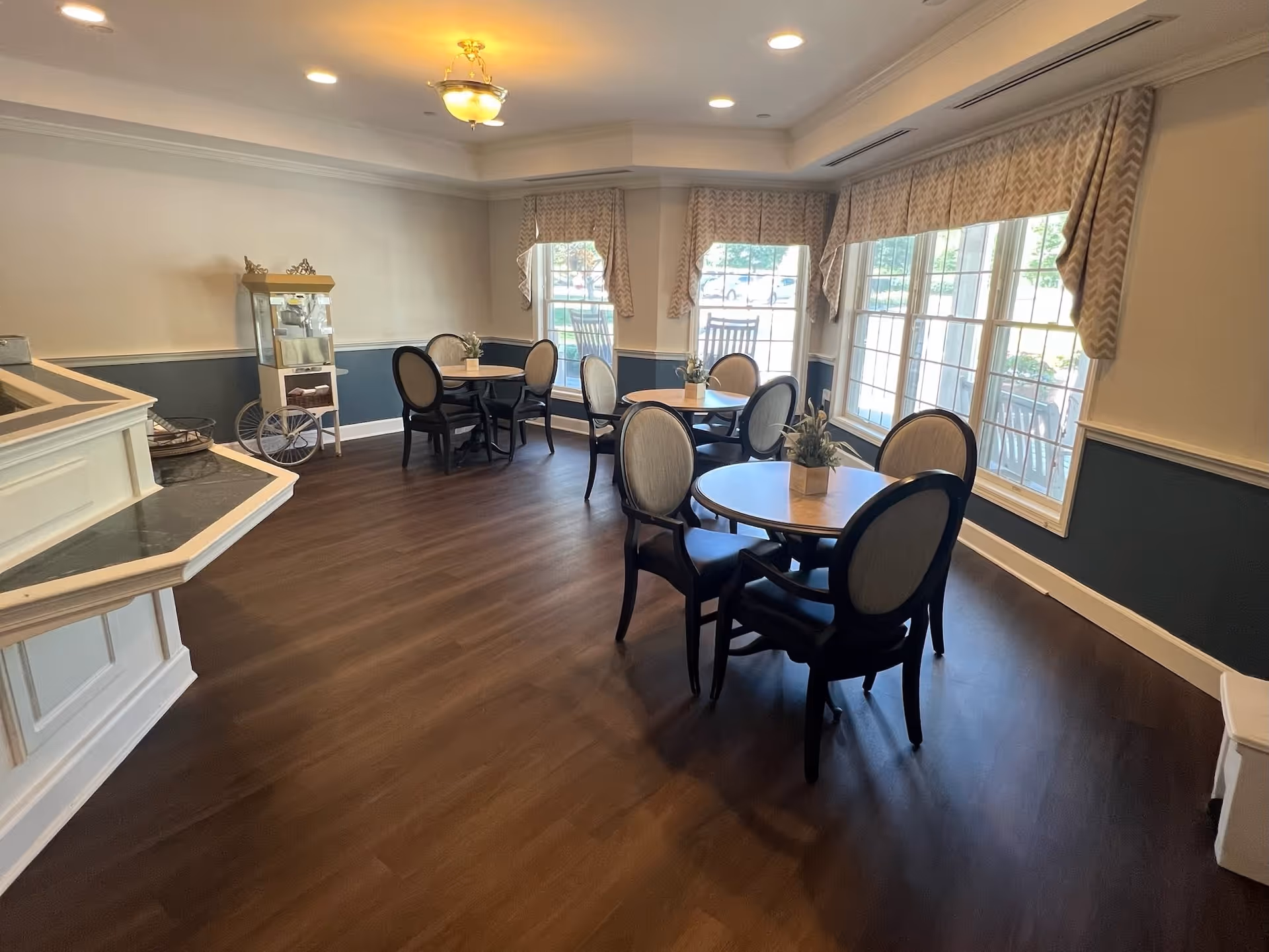 A well-lit dining room with several round tables, each surrounded by four upholstered chairs. The room has large windows with patterned valances, dark wood flooring, and a popcorn machine on a cart in the corner. The walls are painted white on top and dark blue on the bottom with white trim.