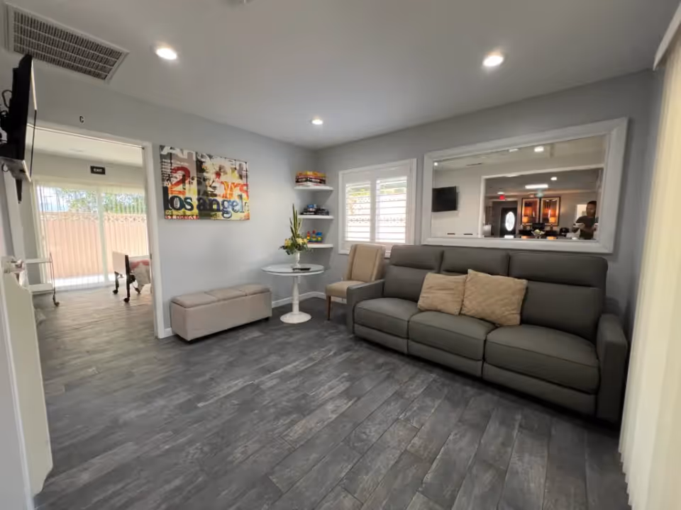 A cozy living room area with a gray sofa adorned with two beige pillows, a single beige chair, and a small round white table with a flower arrangement. The room has gray wood flooring, light gray walls, a large mirror on one wall, and a colorful abstract painting. There is a wall-mounted TV and a doorway leading to another room with a foosball table and an exit door visible.