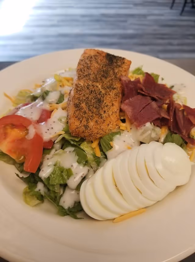 A plate of salad featuring sliced hard-boiled eggs, tomato wedges, shredded cheddar cheese, leafy greens with ranch dressing, a piece of seasoned grilled salmon, and crispy bacon bits.
