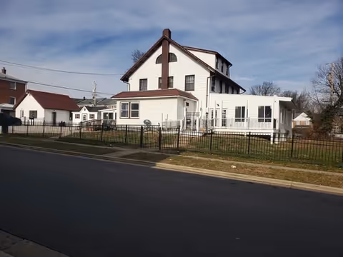 A large white multi-story house with brown trim and a fenced yard, situated along a paved street under a partly cloudy sky.