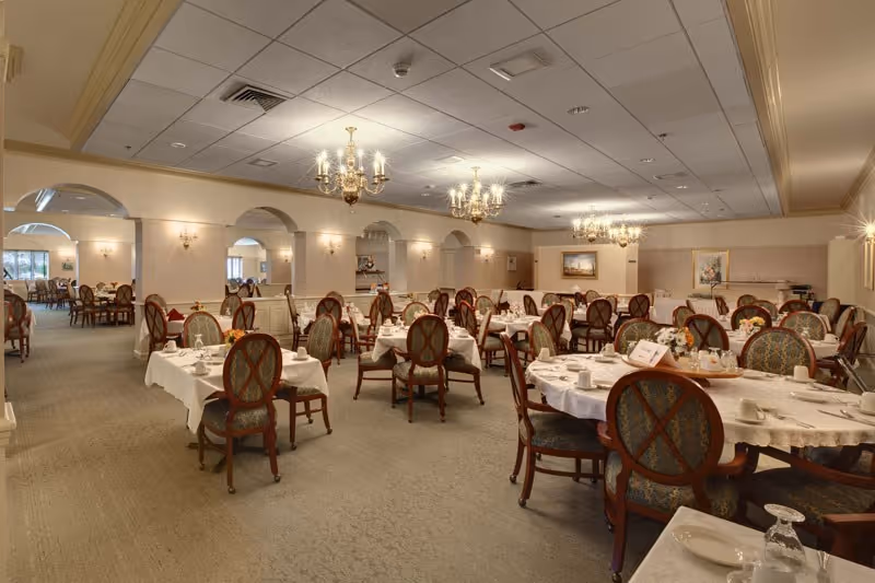 A spacious dining room with multiple round and rectangular tables covered with white tablecloths, set with plates, cups, and silverware. The room features elegant wooden chairs with upholstered seats and backs, chandeliers hanging from the ceiling, and soft lighting from wall sconces. The walls are decorated with framed artwork and mirrors, and there are archways leading to adjacent rooms.