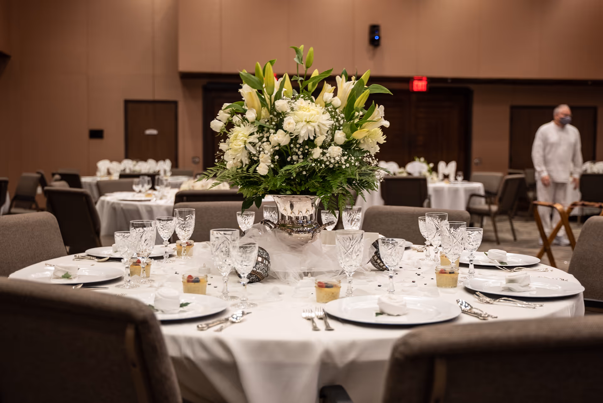 A round dining table set elegantly with white plates, crystal glasses, silverware, and small dessert cups. The centerpiece is a large silver vase filled with white flowers and greenery. The background shows more similarly set tables and a person wearing a mask walking in the room.
