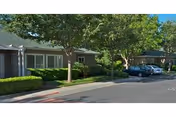 Street view of single-story retirement community cottages with trees, hedges, and parked cars.