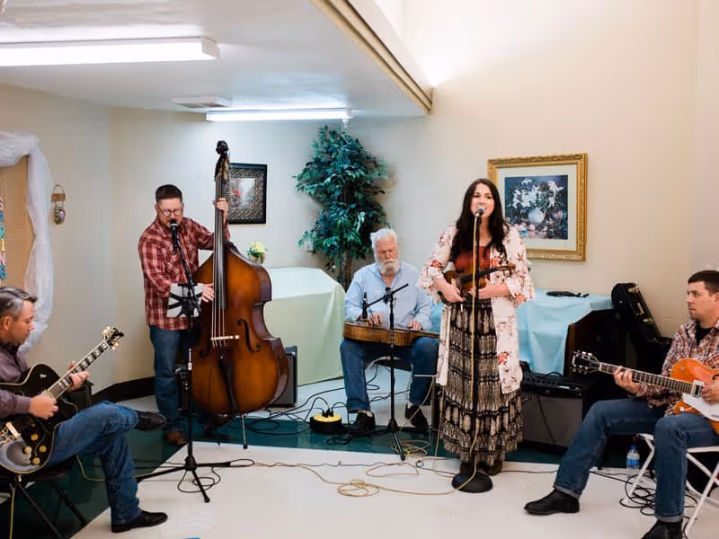 A small musical group performing indoors with five musicians playing instruments including guitars, a double bass, and a hammered dulcimer. One woman is singing into a microphone while holding a violin. The room has beige walls, a framed floral picture, and a potted plant in the background.
