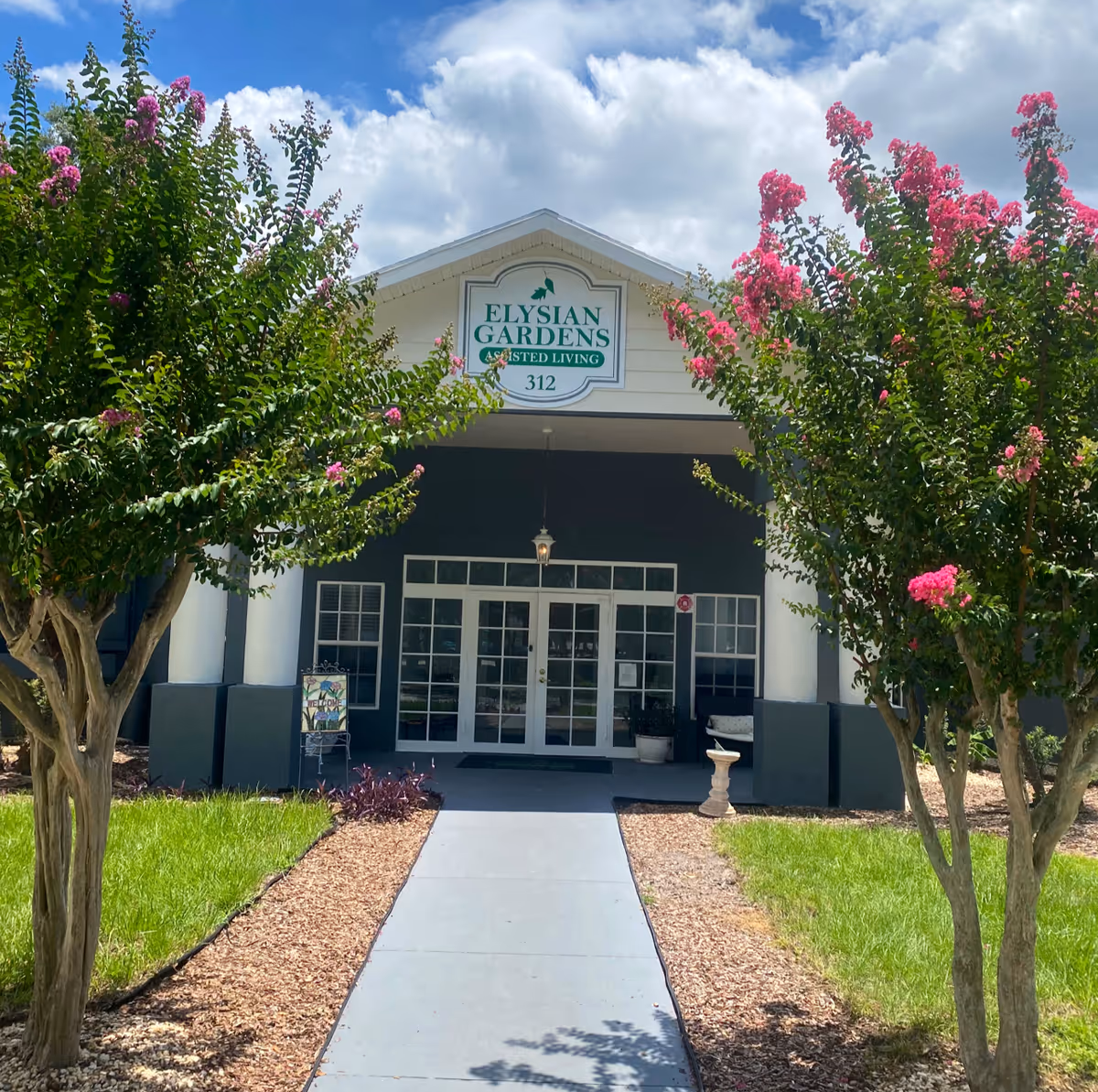 Front entrance of Elysian Gardens Assisted Living Facility with a walkway leading to glass double doors, flanked by two flowering trees with pink blossoms and green grass on either side under a partly cloudy sky.