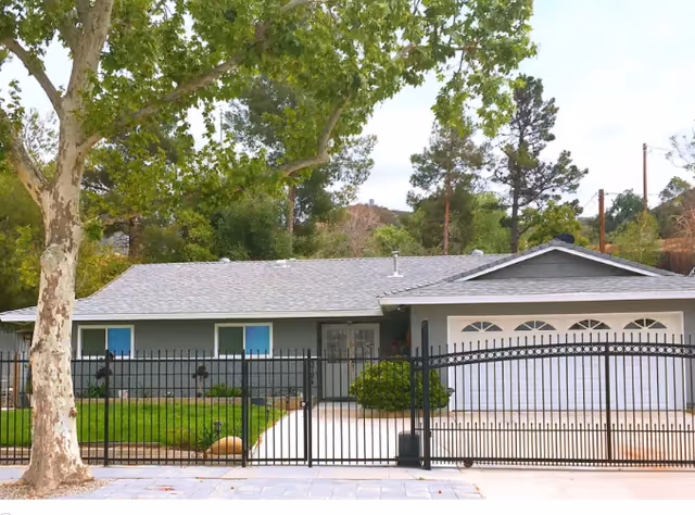 Single-story gray ranch-style house with a gated driveway and attached garage, a front lawn, and a large tree in front.