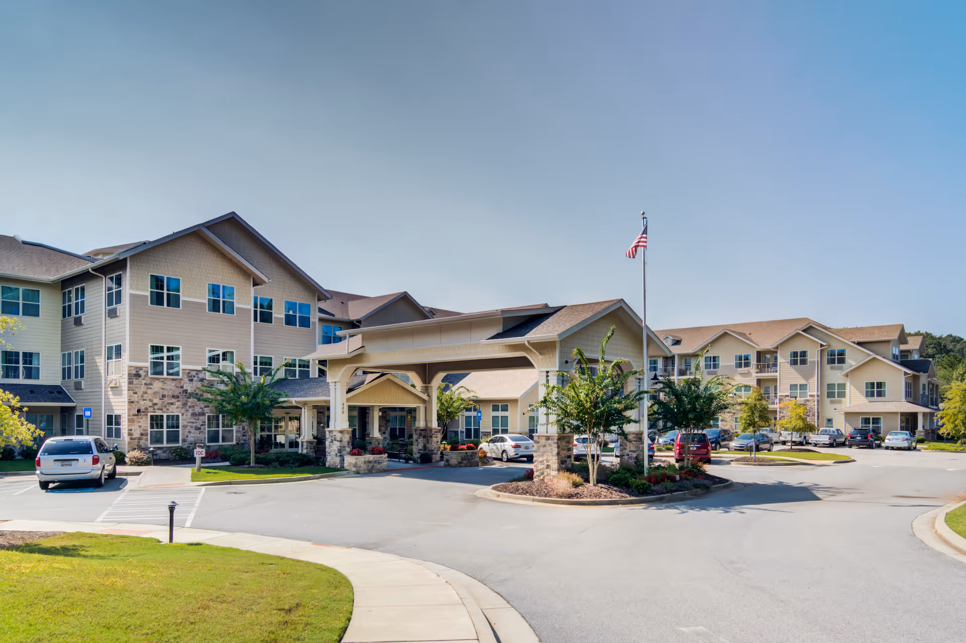 Exterior view of a large, multi-story senior living facility with beige siding and stone accents. The building features many windows and a covered entrance with stone pillars. There is a flagpole with an American flag near the entrance, several parked cars, and landscaped greenery around the driveway.
