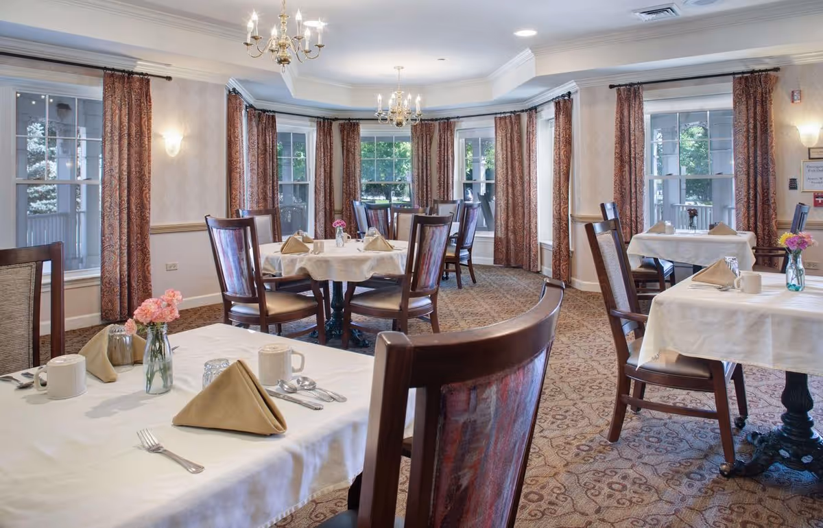 A dining room with multiple tables covered in white tablecloths, each set with beige folded napkins, cups, utensils, and small flower vases. The room has large windows with patterned curtains, carpeted floors, and chandeliers hanging from the ceiling.