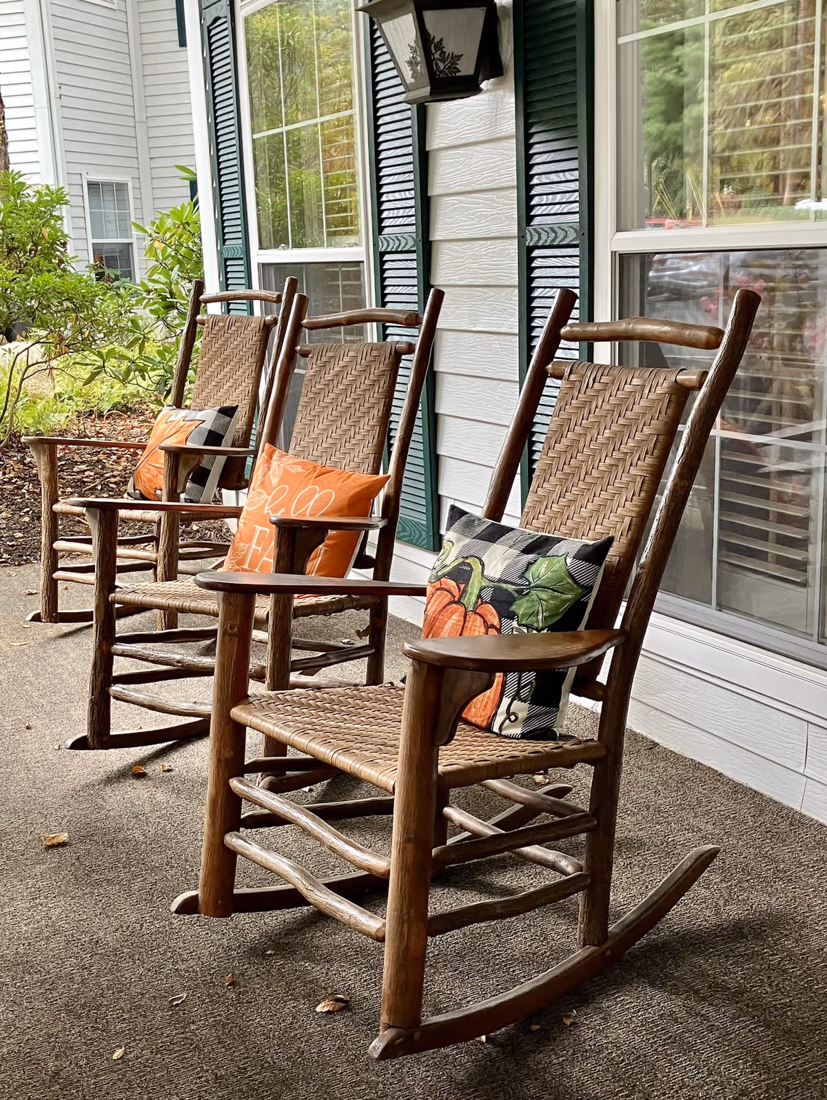 Three wooden rocking chairs with woven seats and backs are arranged on a porch in front of a building with white siding and green shutters. Each chair has a decorative pillow with autumn-themed designs, including pumpkins and fall leaves. There is greenery and a window visible in the background.