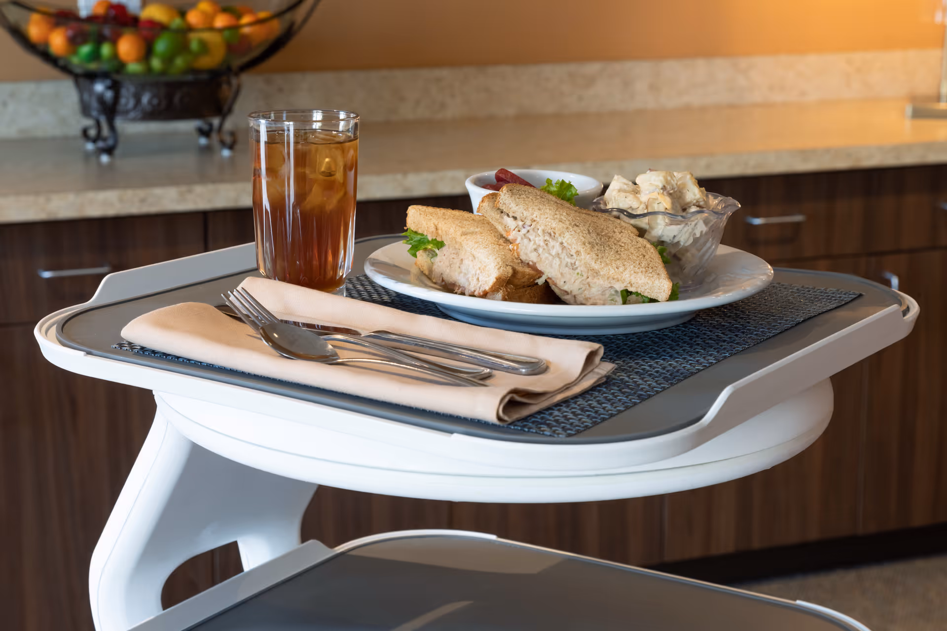 A meal tray on a serving cart holding a sandwich, salad, utensils and a glass of iced tea in a dining area.