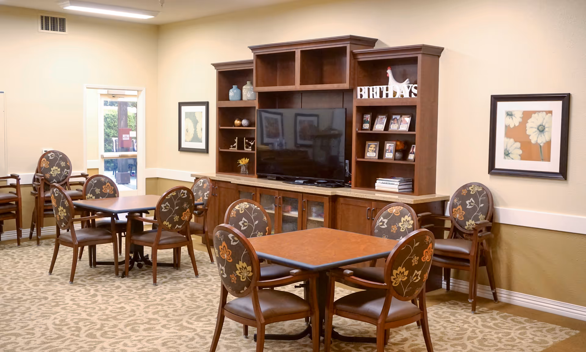 A common area in a senior living facility with multiple square tables surrounded by chairs featuring floral upholstery. A large wooden entertainment center with a flat-screen TV and decorative items is against the wall. The room has beige walls, patterned carpet, framed floral artwork, and a door leading outside.