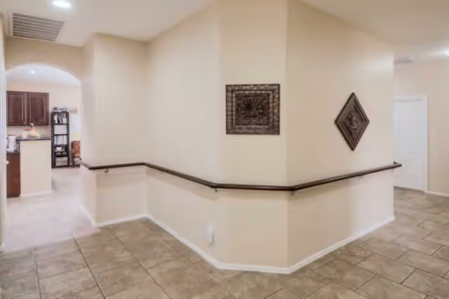 Interior hallway with beige walls, tile floor, wall-mounted handrails and decorative wall plaques opening toward a kitchen area.