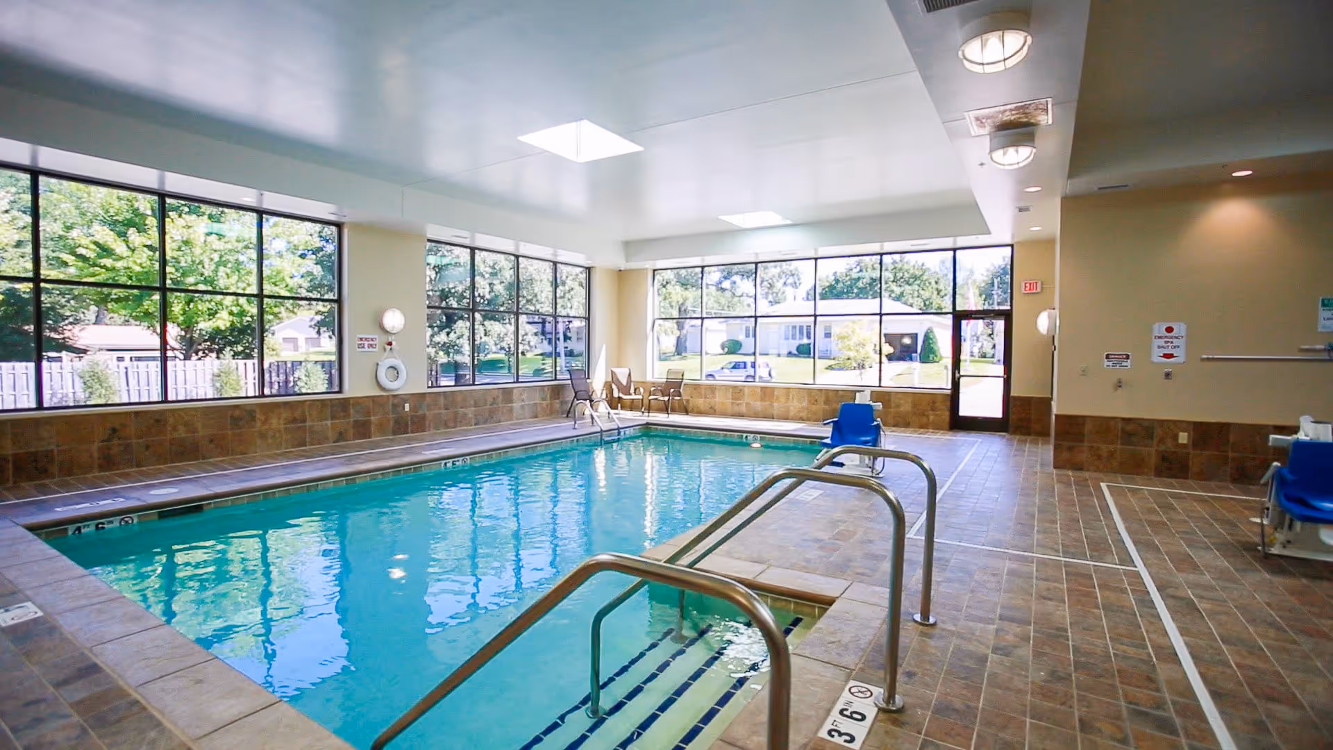 Indoor swimming pool area with large windows letting in natural light, tiled floors and walls, metal handrails for pool access, and a pool lift chair for accessibility. Outside greenery is visible through the windows.
