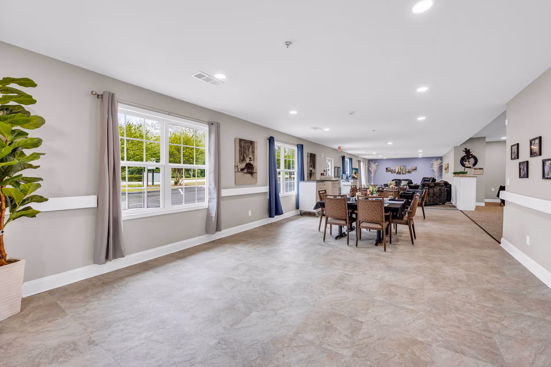 A spacious, well-lit common area in a senior living facility with large windows on the left side, beige walls, and light-colored flooring. There are several tables with chairs arranged for dining or activities, and a seating area with sofas and armchairs in the background. The room is decorated with framed pictures on the walls and potted plants.