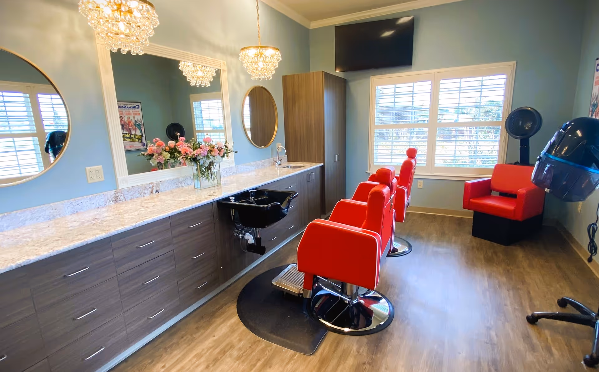 Interior of a salon room with three red salon chairs, a black hair washing sink, two round mirrors, a large rectangular mirror, chandeliers, a window with white blinds, and a red drying chair with a hooded hair dryer.