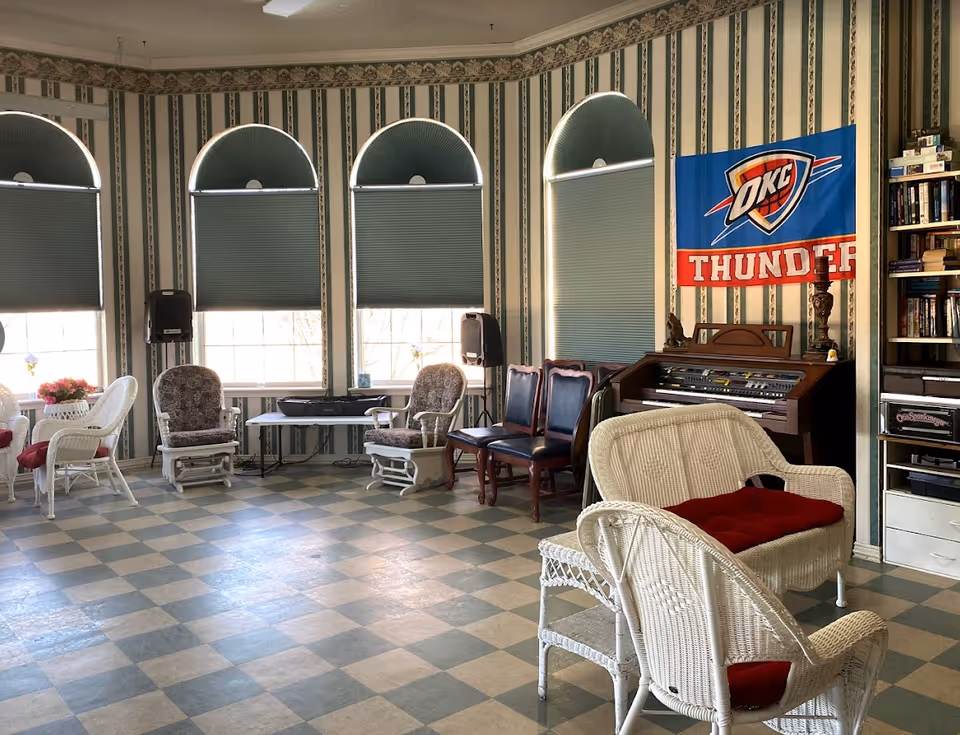 Sunlit common room with wicker chairs and cushioned armchairs arranged near arched windows, an organ and an OKC Thunder banner on the wall.