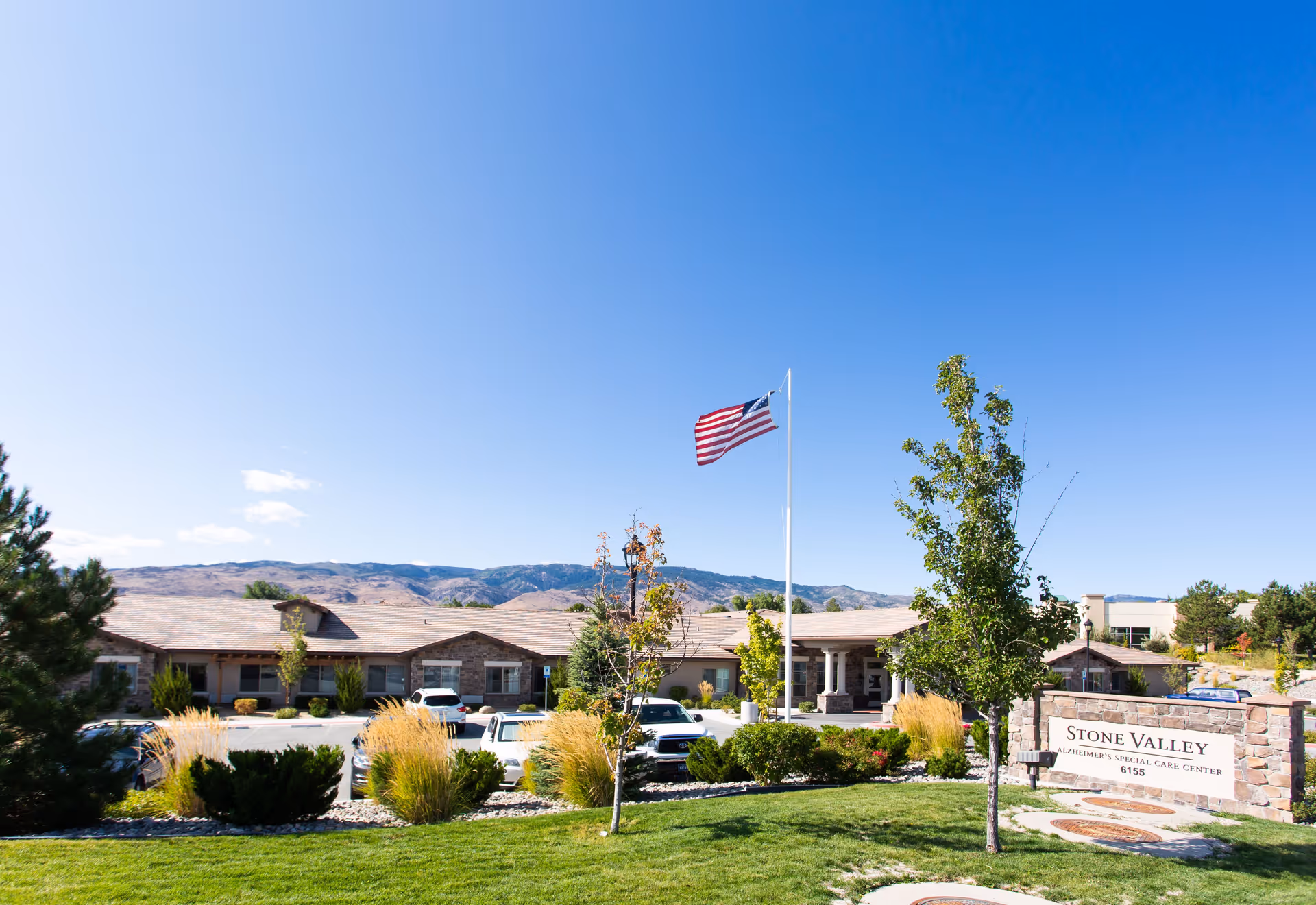 Exterior view of Stone Valley Assisted Living & Memory Care facility with a clear blue sky, an American flag on a flagpole, landscaped greenery, and a parking area with several cars in front of the building.