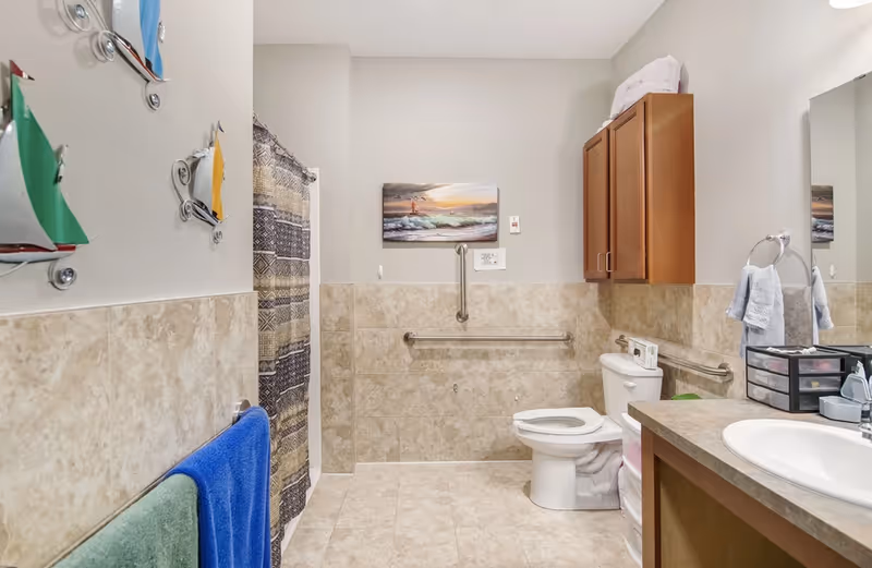 Accessible bathroom with toilet, sink and countertop, shower with curtain, grab bars, towels and wall cabinets.