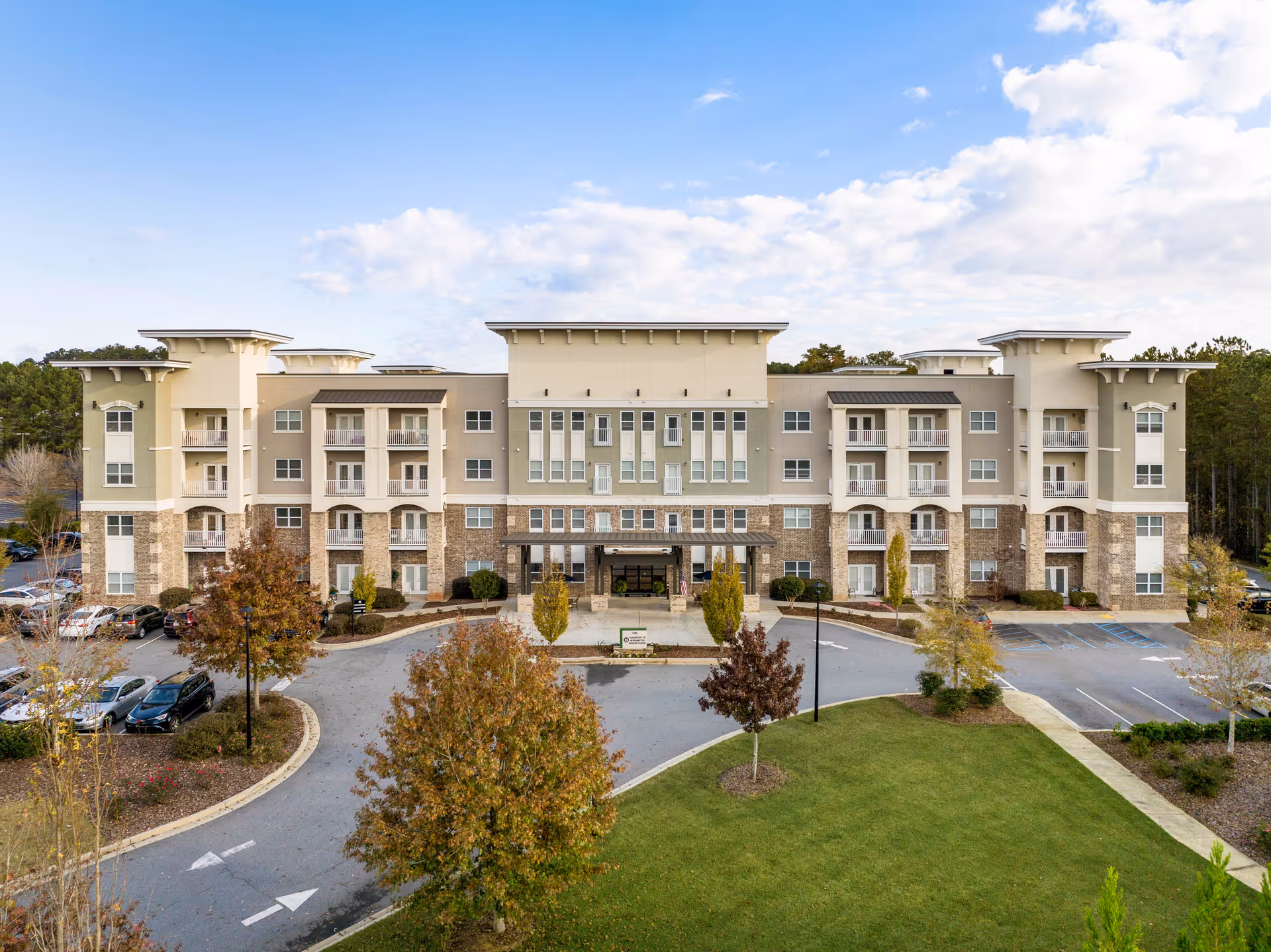 Front exterior of a four-story beige senior living building with balconies, a covered entrance drive, parked cars, and landscaped grounds.
