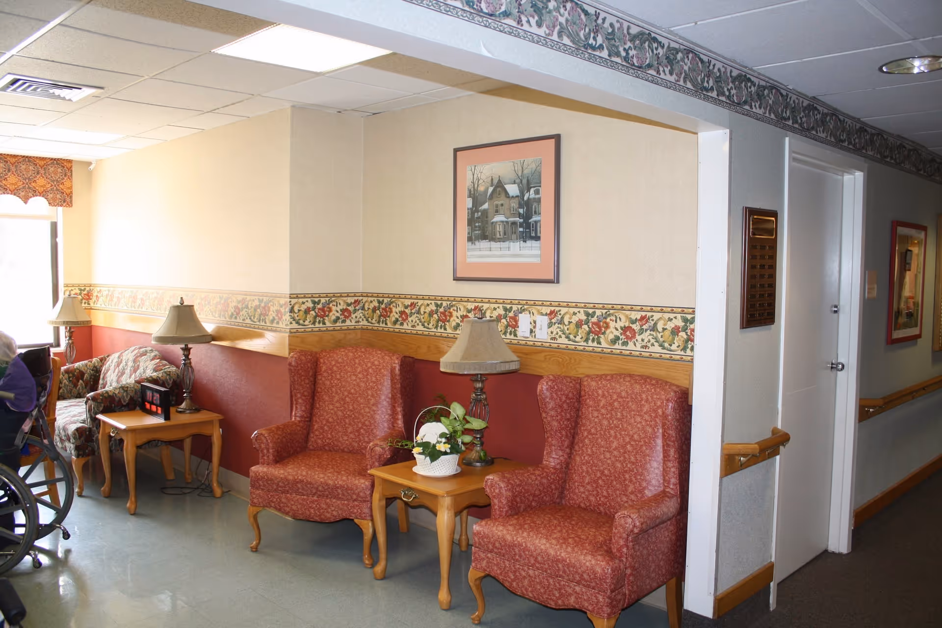 A cozy seating area in a senior living facility with two red patterned armchairs and a wooden side table with a lamp and a potted plant. Another armchair and side table with a lamp are visible near a window with a floral valance. The walls have a floral wallpaper border and framed artwork. A hallway with handrails and doors is visible to the right.