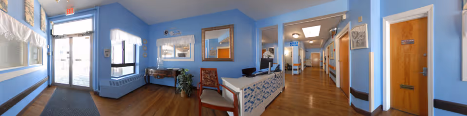 Interior view of a healthcare facility reception area with blue walls and wooden flooring. There is a reception desk with a computer and chair, a decorative table with a plant, and framed artwork on the walls. A hallway extends from the reception area with doors on either side and ceiling lights. A glass exit door is visible on the left side with natural light coming through.