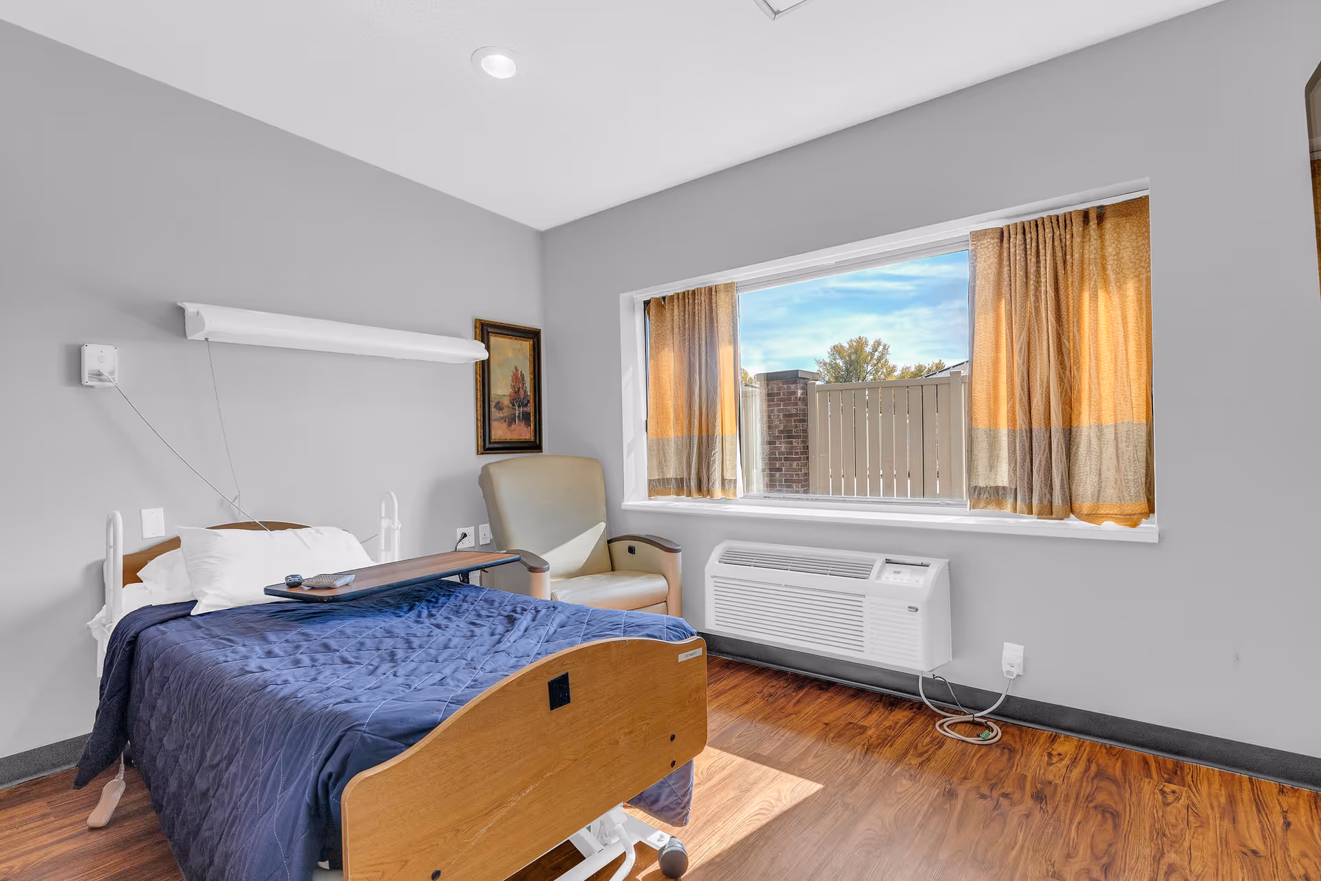 A private senior living bedroom with an adjustable single bed, overbed table, armchair, large window with curtains, and a wall-mounted air conditioner.