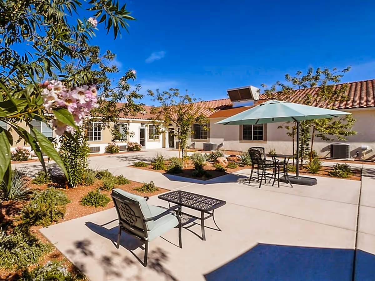 Outdoor courtyard area of Silver Hills Health Care Center featuring a concrete patio with a cushioned chair, a metal table, and a seating area with a table and umbrella. Surrounding the patio are landscaped garden beds with small trees, bushes, and flowering plants under a clear blue sky.