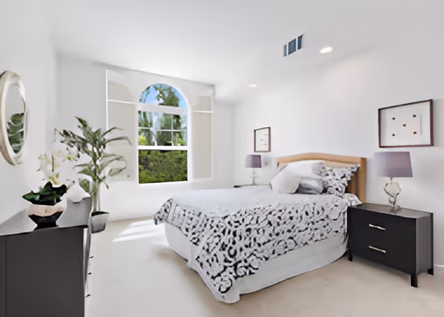 Bright bedroom with a bed, two nightstands, a dresser, potted plants, and an arched window showing greenery.