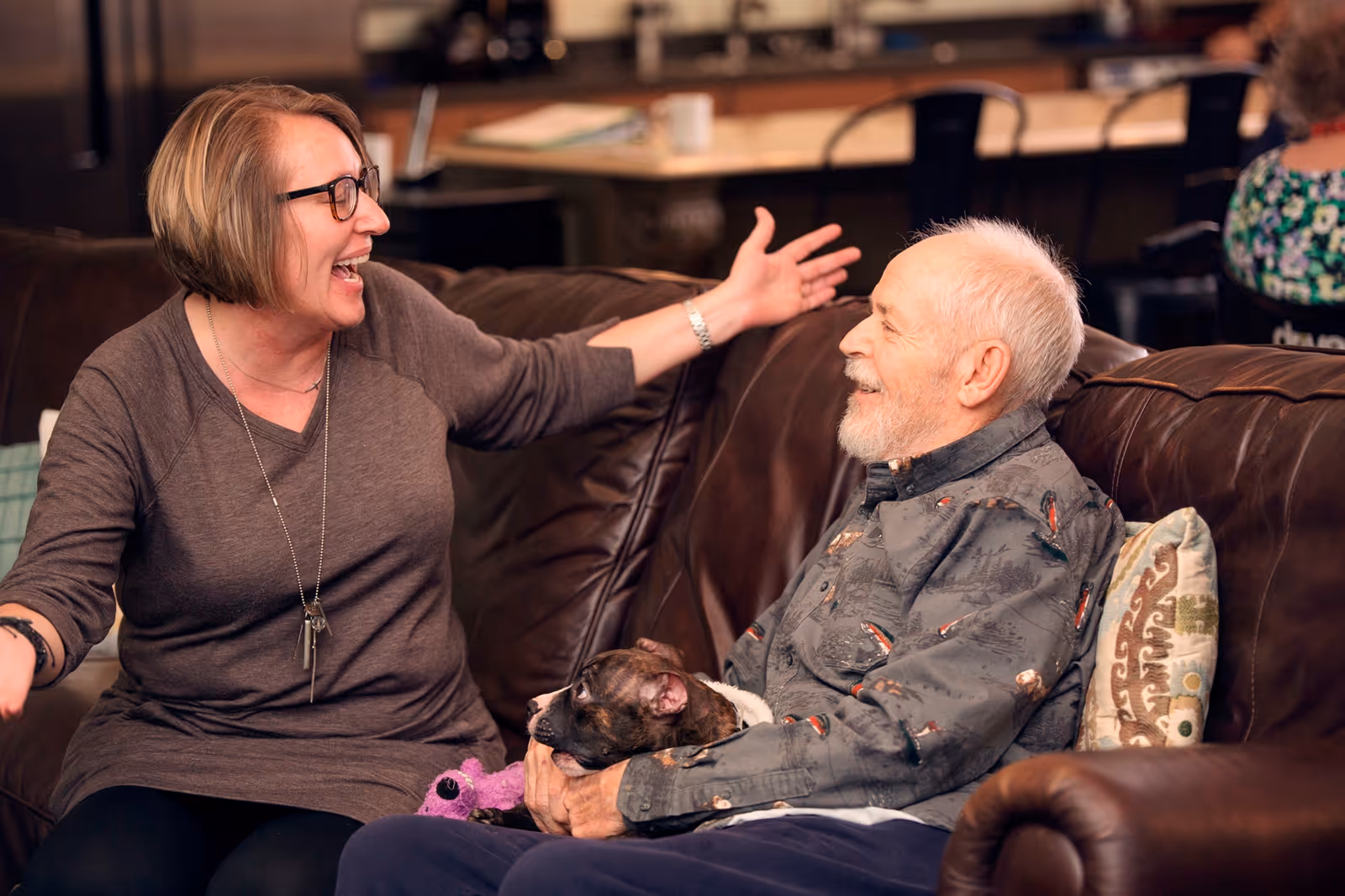 An elderly man sitting on a brown leather couch holding a small dog, smiling and looking at a woman sitting next to him who is laughing and gesturing with her arms open. The setting appears to be a cozy living room or common area.