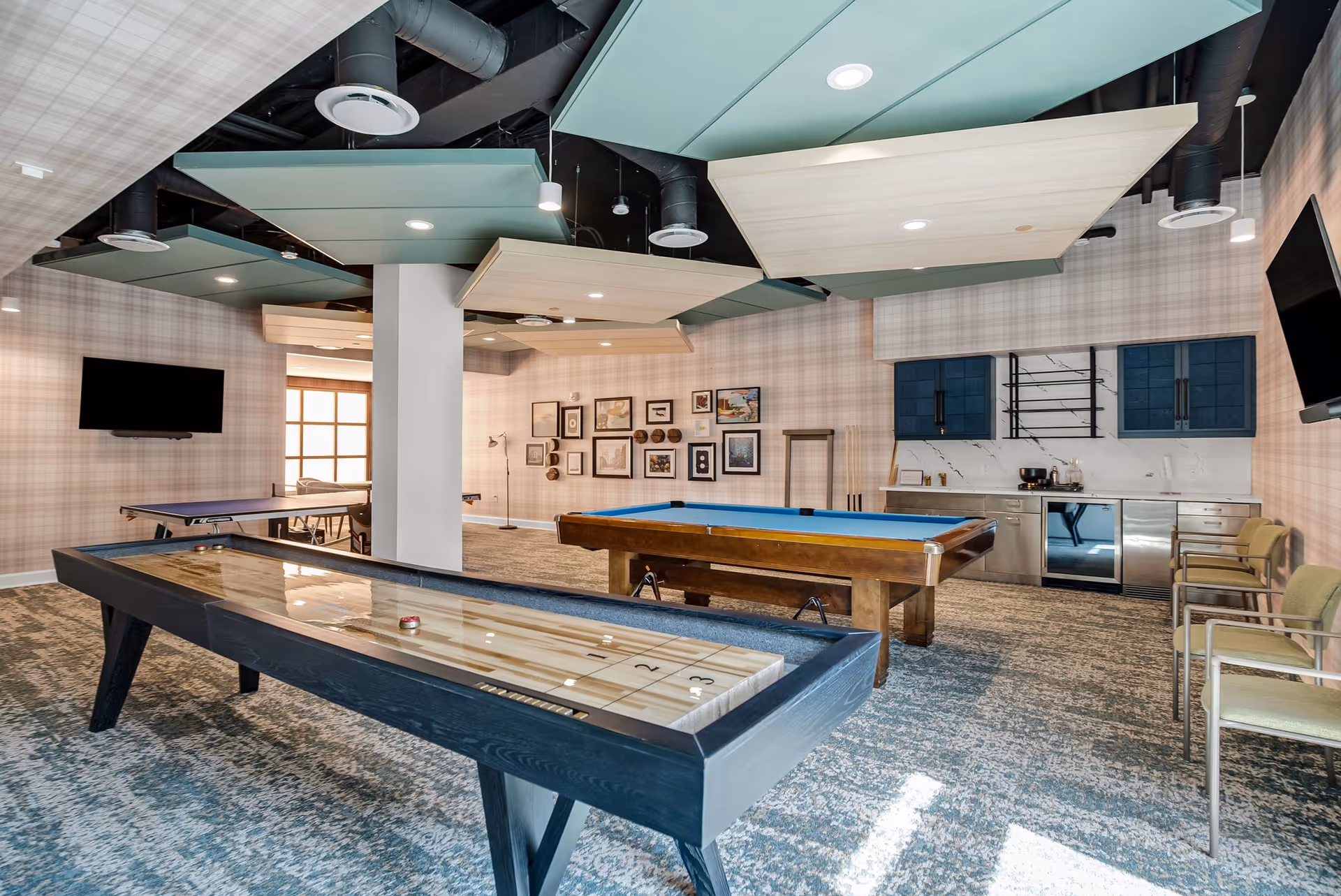 Recreation room with shuffleboard and pool tables, a kitchenette, seating, wall art, and mounted TVs.
