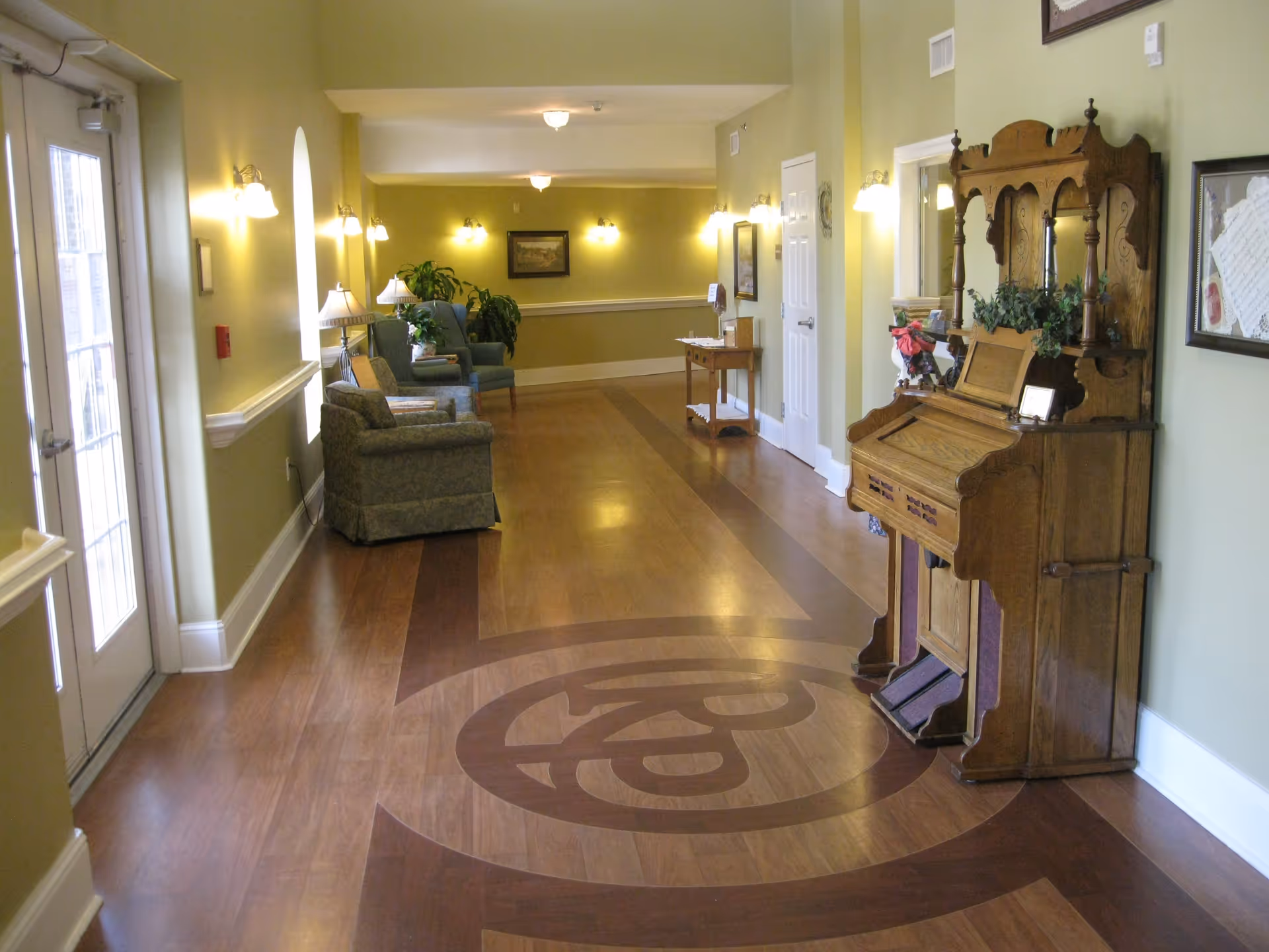 A hallway in a senior living facility with light green walls and wooden flooring featuring a decorative circular pattern. The hallway is furnished with upholstered armchairs, small tables with lamps, plants, framed artwork on the walls, and a vintage wooden organ. Wall-mounted lights illuminate the space, and a glass door is visible on the left side.