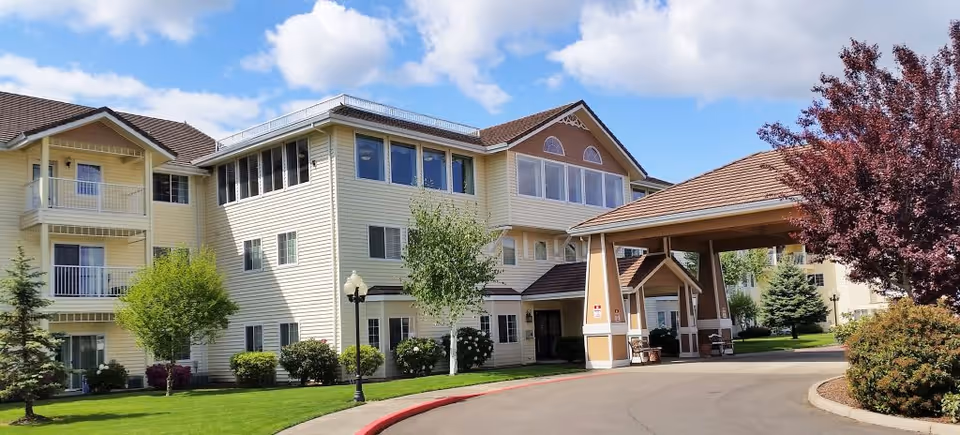 Exterior view of a multi-story senior living facility with light yellow siding, multiple windows, and a covered entrance driveway. The building is surrounded by well-maintained green lawns, trees, and shrubs under a partly cloudy blue sky.