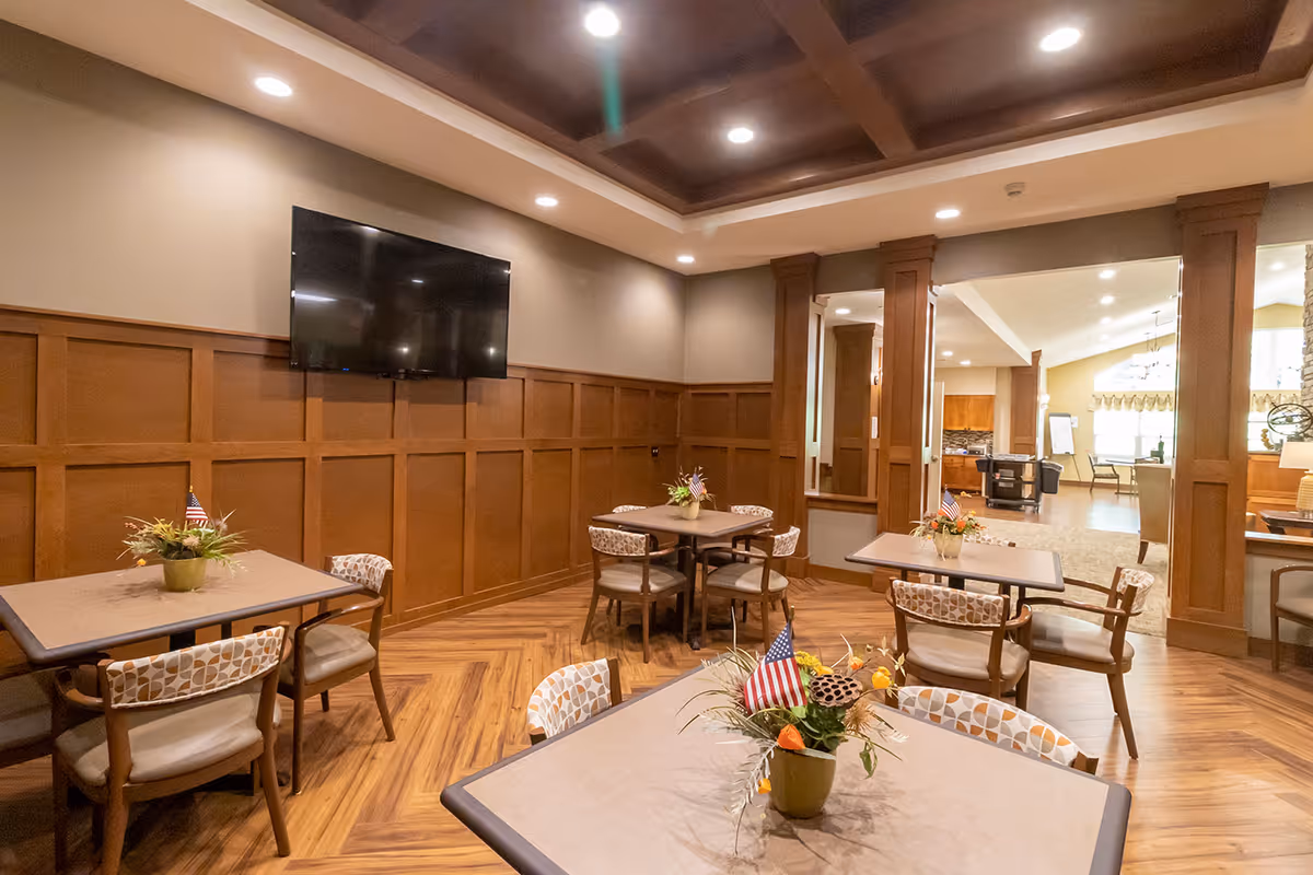 Cozy dining/activity room with tables and chairs, wood-paneled walls, a wall-mounted TV and small floral centerpieces.