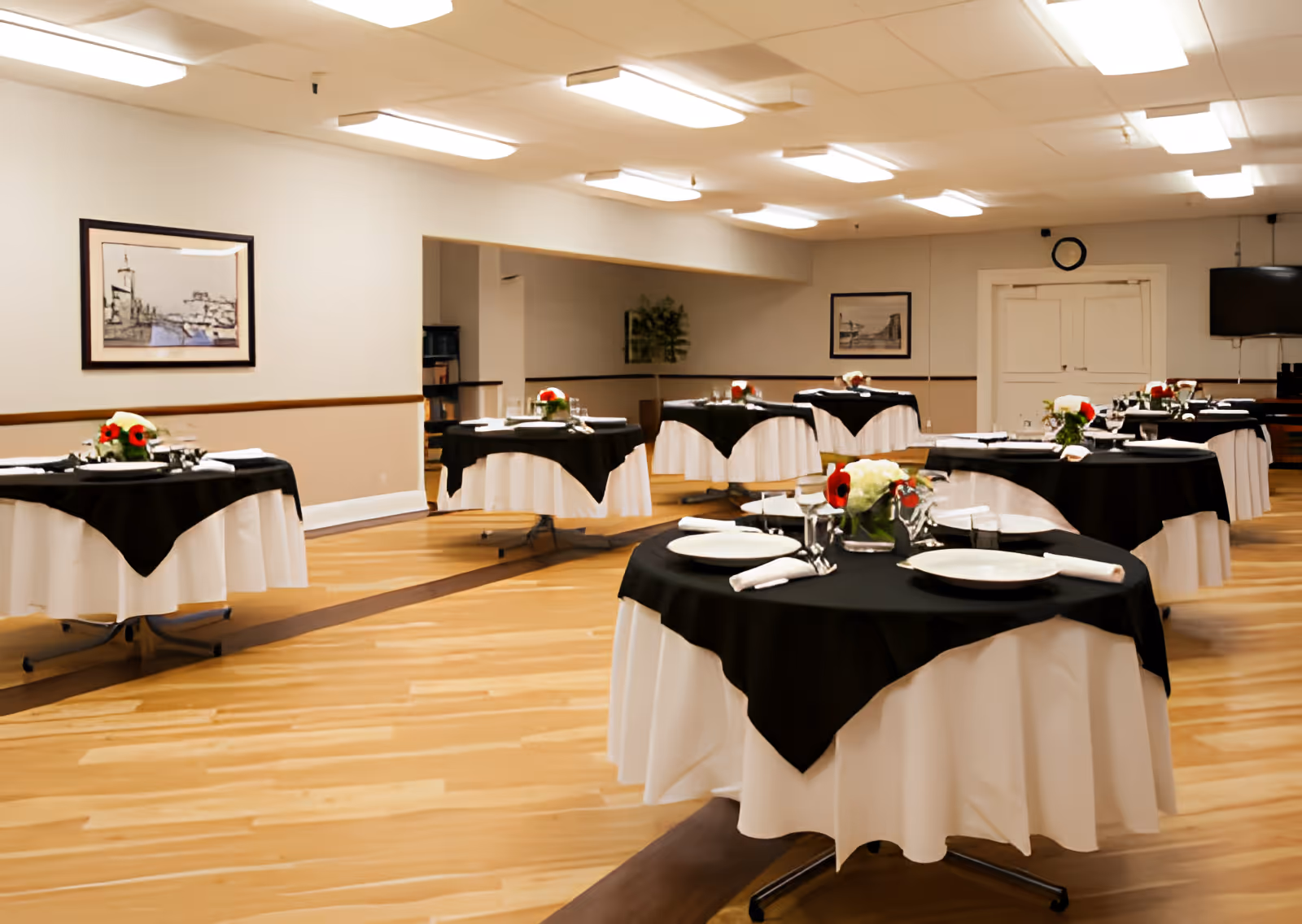 A dining room with round tables covered in white tablecloths and black overlays, each set with plates, napkins, glasses, and floral centerpieces. The room has wooden flooring, white walls with framed artwork, and bright ceiling lights.
