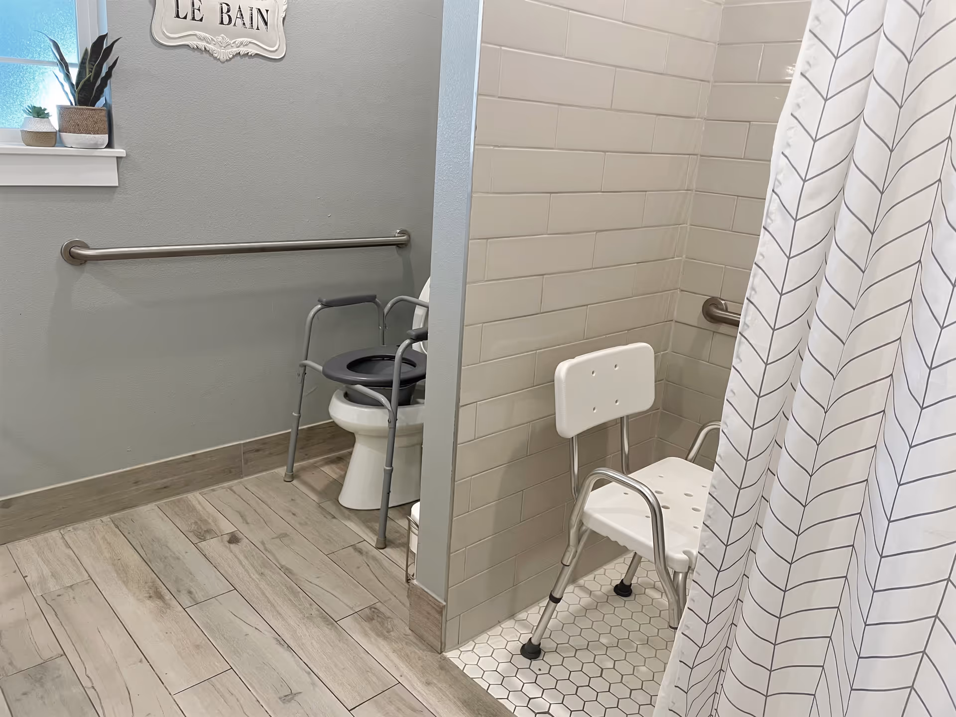 Accessible bathroom with a toilet equipped with a raised seat and armrests, a grab bar on the wall, a small window with potted plants on the sill, and a shower area with a white shower chair and a curtain with a leaf pattern.