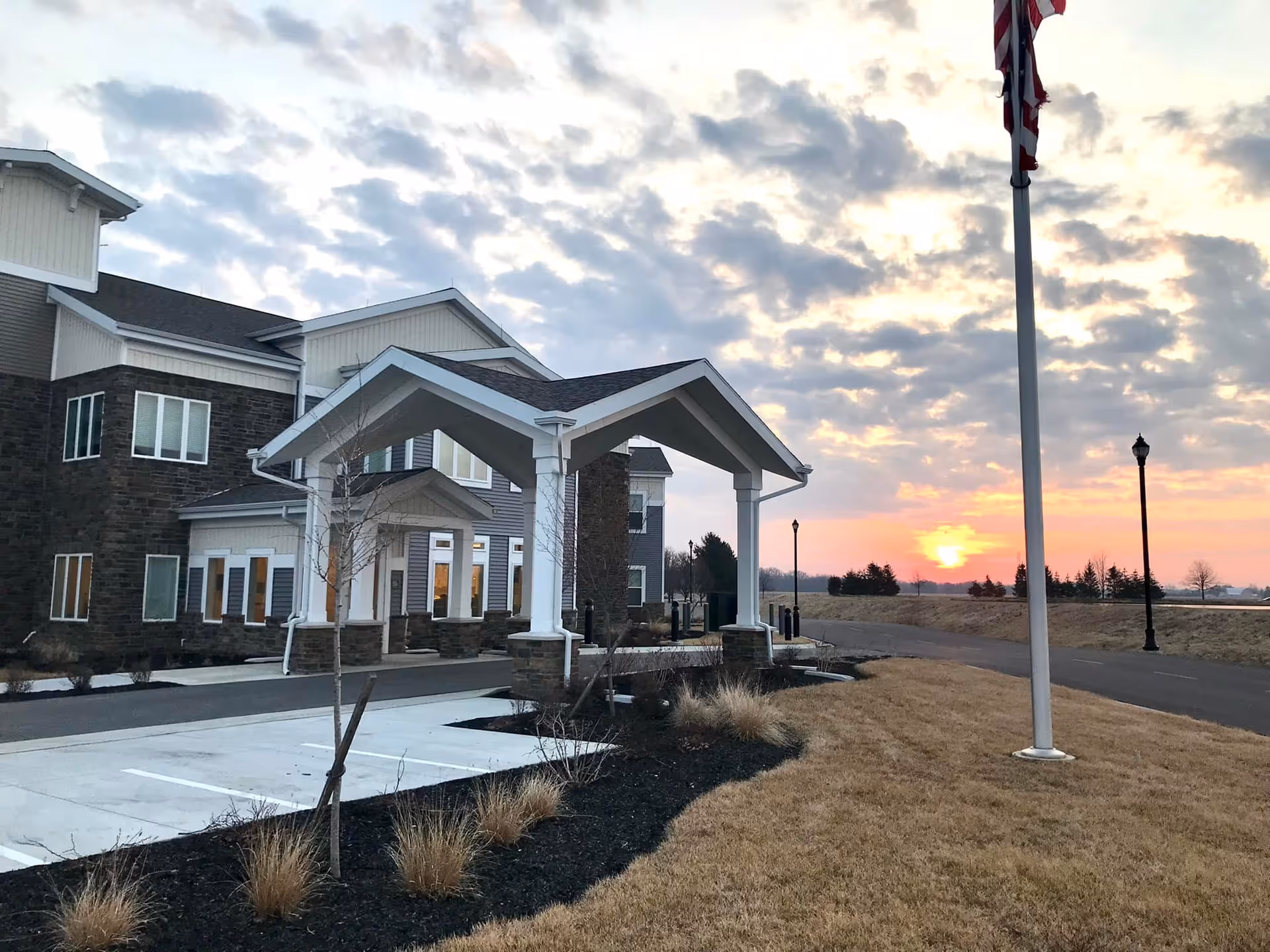 Exterior view of Otterbein Pemberville SeniorLife Community building at sunset with a covered entrance, landscaped grounds, and an American flag on a flagpole.