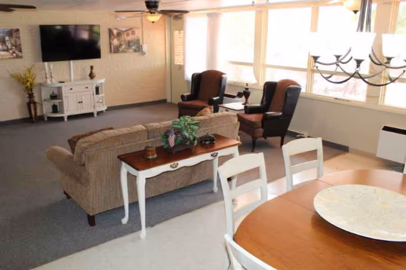 A bright living room area with a beige couch, two brown armchairs, a white console table with decorative items, a wall-mounted flat screen TV, and a dining table with white chairs in the foreground. Large windows allow natural light to fill the space.