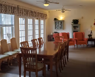 A spacious dining room with a long wooden table surrounded by chairs. Large windows with floral valances let in natural light. In the background, there are two red armchairs, a small cabinet, and a side table with a lamp and plant. Ceiling fans and wall sconces provide lighting.