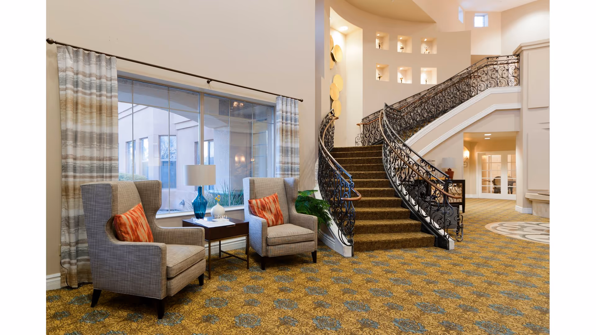 A spacious and elegant interior common area featuring a curved staircase with ornate black railings. Two gray upholstered armchairs with orange patterned pillows are positioned near a large window with striped curtains. A small table with a blue glass lamp and decorative items sits between the chairs. The carpet has a yellow and blue pattern, and the walls are painted in a light neutral color.